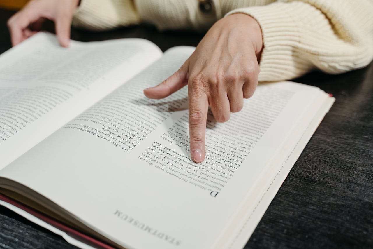 Close-up of a hand pointing at text in an open book on a table.