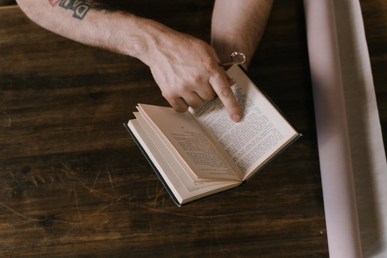 Close-up of a hand pointing at text in an open book on a wooden table.